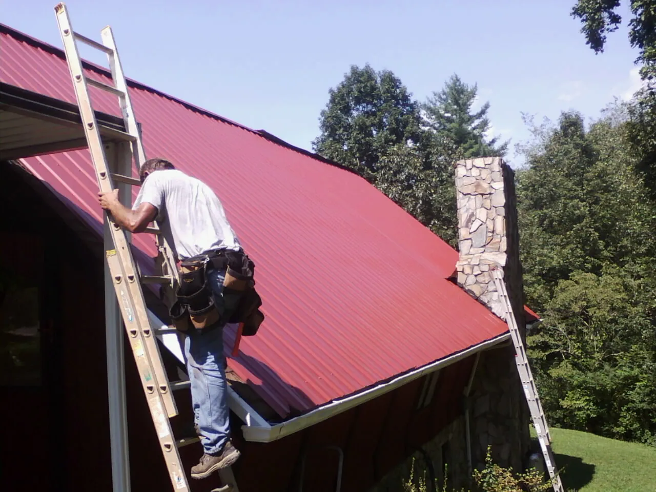 Roofer on a ladder beside a red metal roof and stone chimney — Soffit & Fascia Repair by Roofing Brothers in Rex